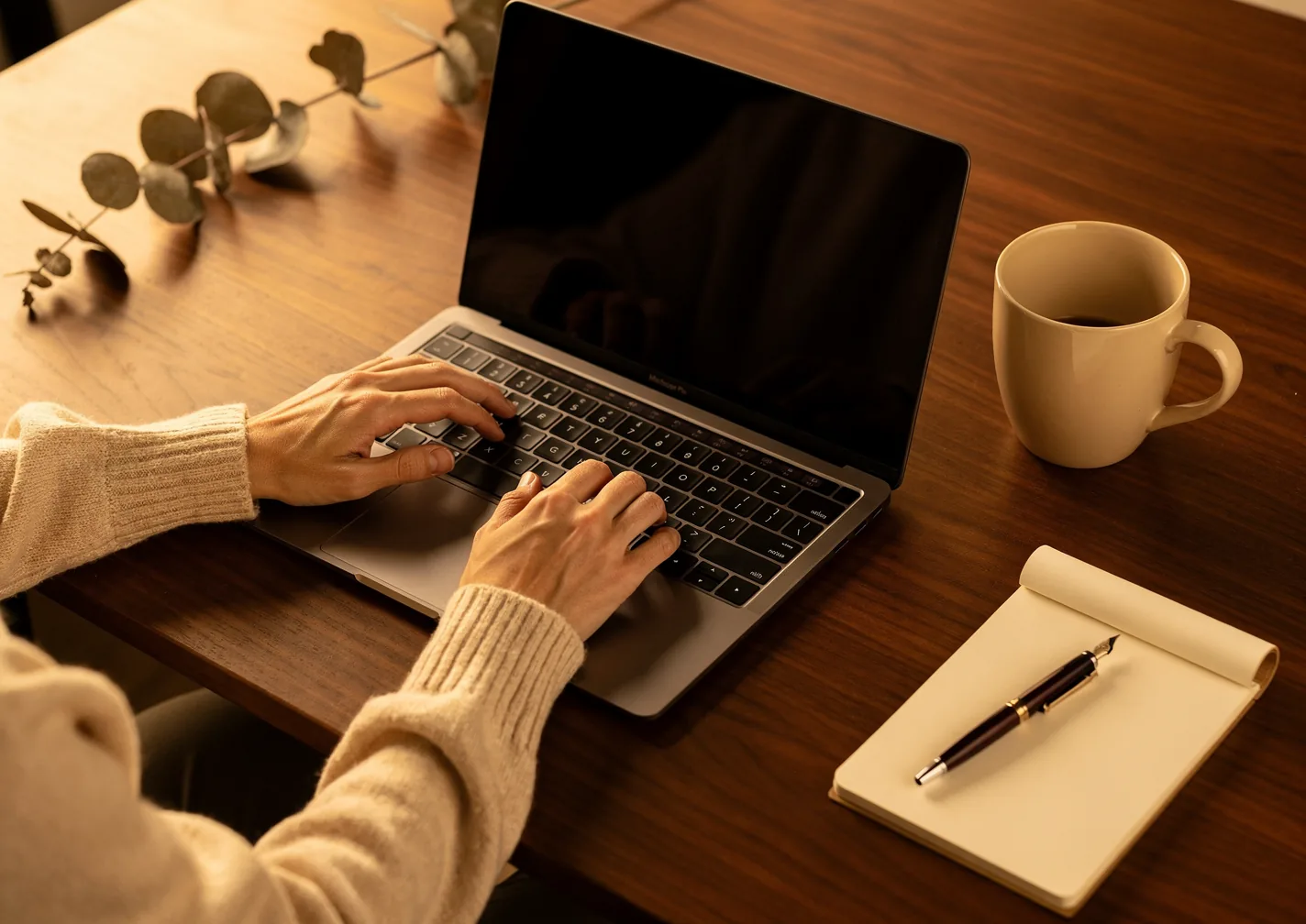 Des mains tapent sur un ordinateur portable, une tasse et un carnet à côté